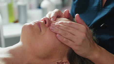 high angle close up of skin care expert placing hands on reclined woman on massage bed and gently massaging eyelid and surrounding eye area with smooth strokes under bright spa lighting
