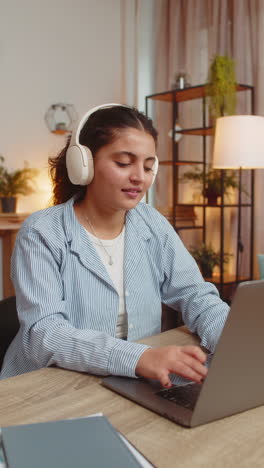 happy indian woman using laptop while listening to music via headphones sitting at home office desk