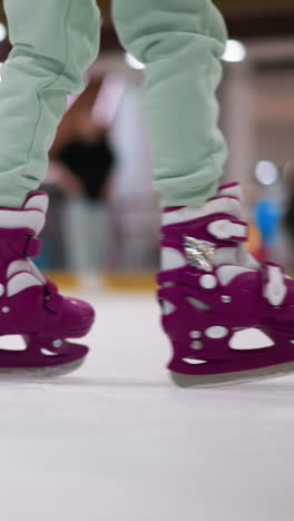 primer plano de alguien patinando en patines de hielo púrpura en una pista de hielo, con pantalones de color ceniza, con otras personas patinando al fondo y una atmósfera animada