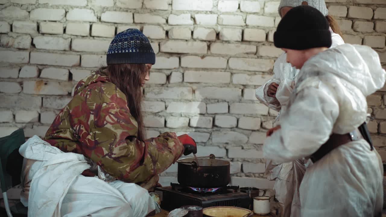 Survivor in worn camouflage jacket and knitted hat places black pot on stove inside cold brick shelter, children gather nearby to drop herbs into boiling food while scene reflects harsh struggle