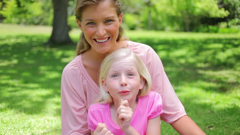 a woman sits behind her daughter who is repeatedly blowing a dandelion before smiling