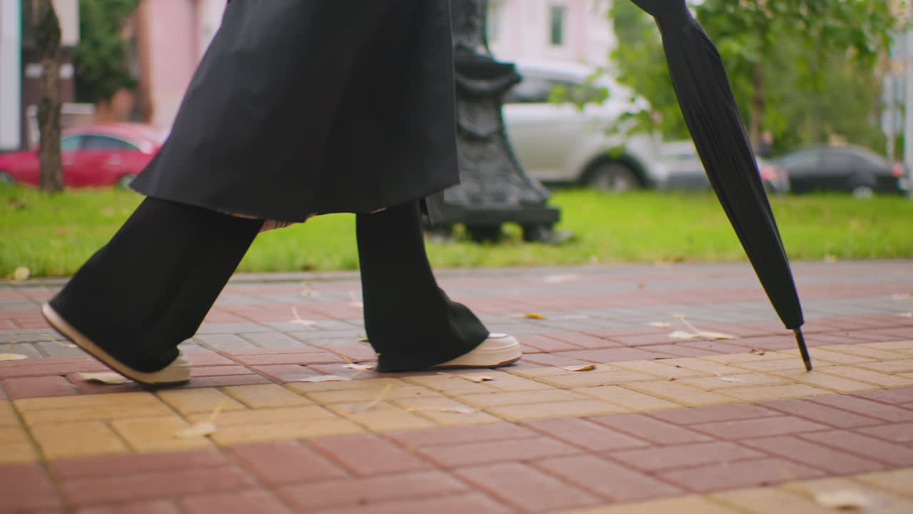 Close up of woman walking on colorful pavement wearing black trench coat and white sneakers, holding closed umbrella, urban background with cars and greenery, autumn atmosphere with scattered leaves