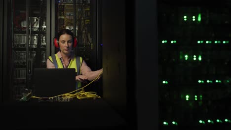 caucasian female it technician with headphones on using laptop and checking computer server