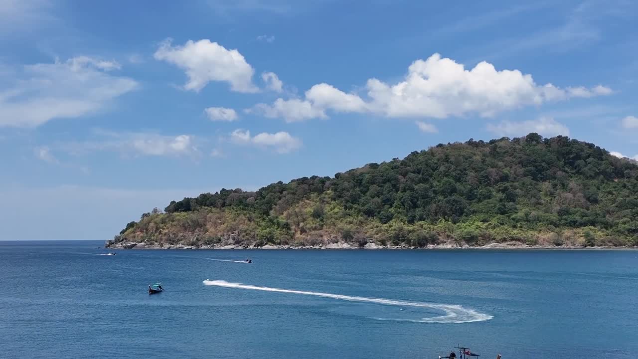 드론 영상은 밝고 이 많은 하늘 아래 푸켓의 바위 해안선을 따라 은 물을 항해하는 롱테일 보트를 포착합니다.