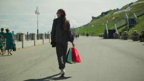dama con ropa gris llevando bolsas de compras rojas, menta y verdes, camina por un puente con postes de luz a un lado y una colina al otro, madre con hijos paseando cerca del borde del puente