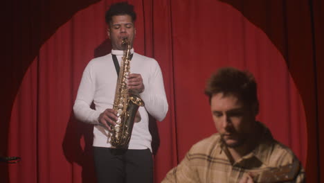 latin man playing sax and man sitting and playing guitar on stage during a rehearsal