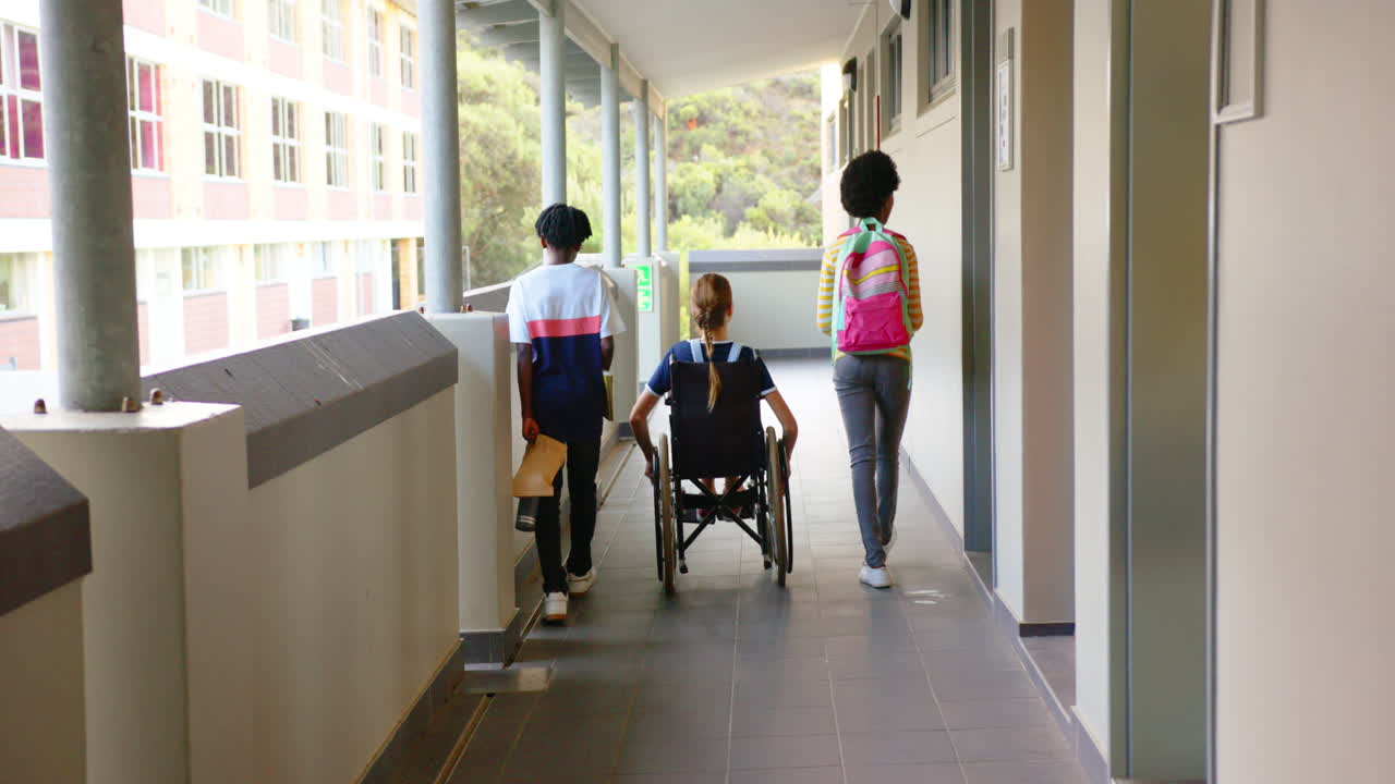 Walking in school hallway, girl students with backpacks accompanying friend in wheelchair