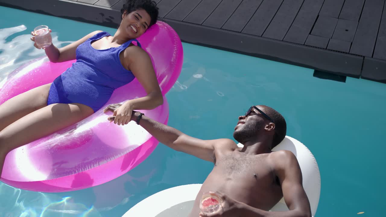 Diverse couple noticing each other, reaching across pink floats, passing pink drink at private pool