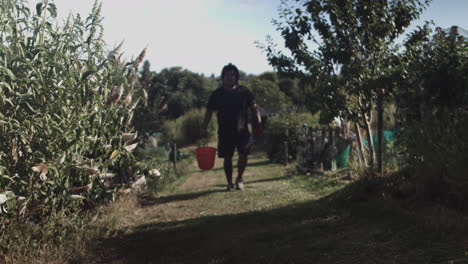 Full-shot-of-a-latin-man-walking-forward-through-his-yard-with-gloves-and-an-orange-bucket-at-daytime