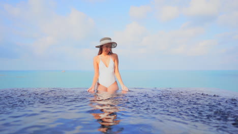 Pretty-female-tourist-sitting-on-the-edge-of-infinity-pool,-having-fun-and-splashing-the-water-with-her-arms-,-with-the-ocean-in-the-background-during-bright-sunny-day