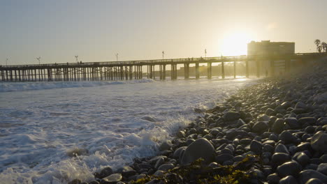 Tiro-De-Muñeca-Avanzando-A-Lo-Largo-De-La-Costa-Rocosa-De-Una-Playa-Del-Sur-De-California-Mientras-Las-Olas-Se-Estrellan-Con-El-Muelle-De-Riesgo-En-El-Fondo-Ubicado-En-El-Sur-De-California