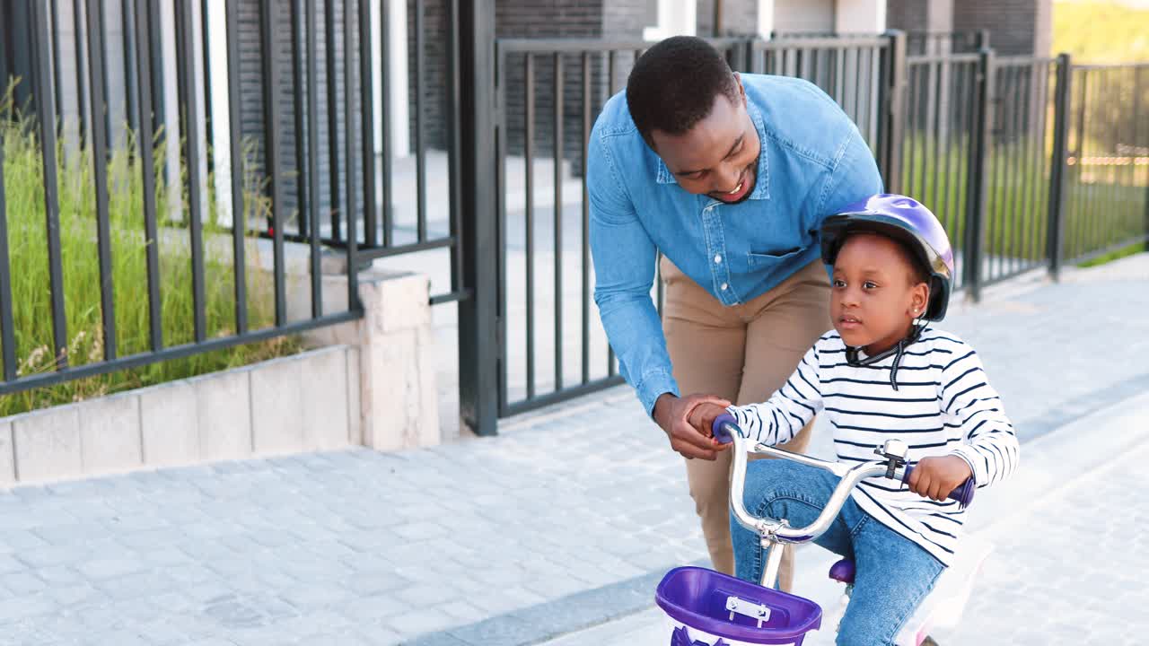 African American father teaching small girl in helmet riding on bike at street in outskirt