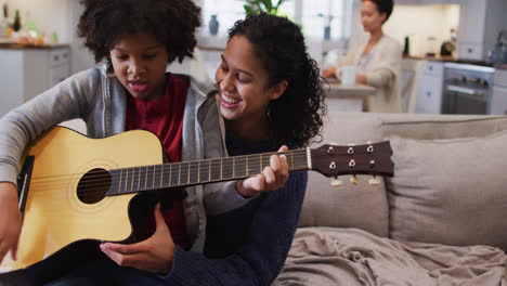 Mixed-race-mother-and-daughter-sitting-on-couch-playing-guitar