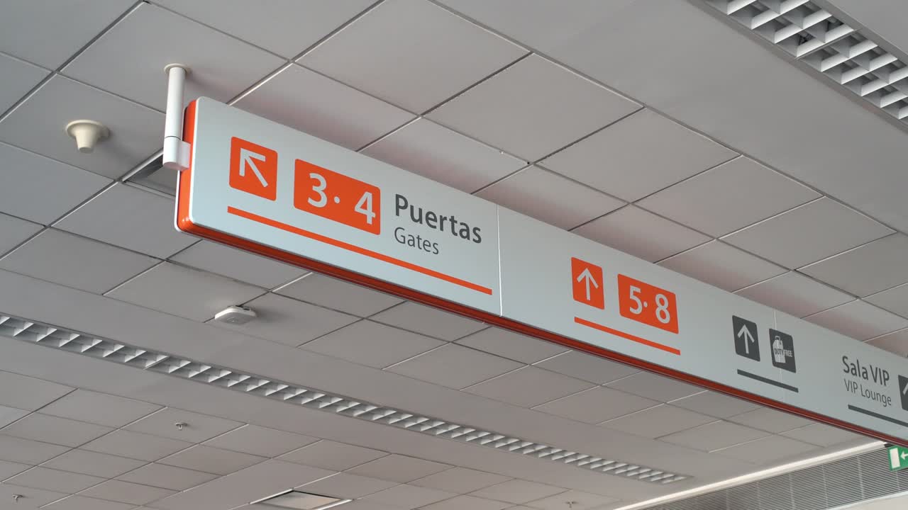 Señal direccional bilingüe colgando del techo de una terminal de aeropuerto moderna que indica puertas de embarque, ascensores y la sala VIP