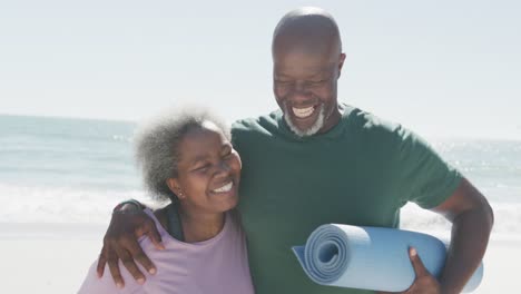 happy senior african american couple embracing and holding yoga mats at beach, in slow motion