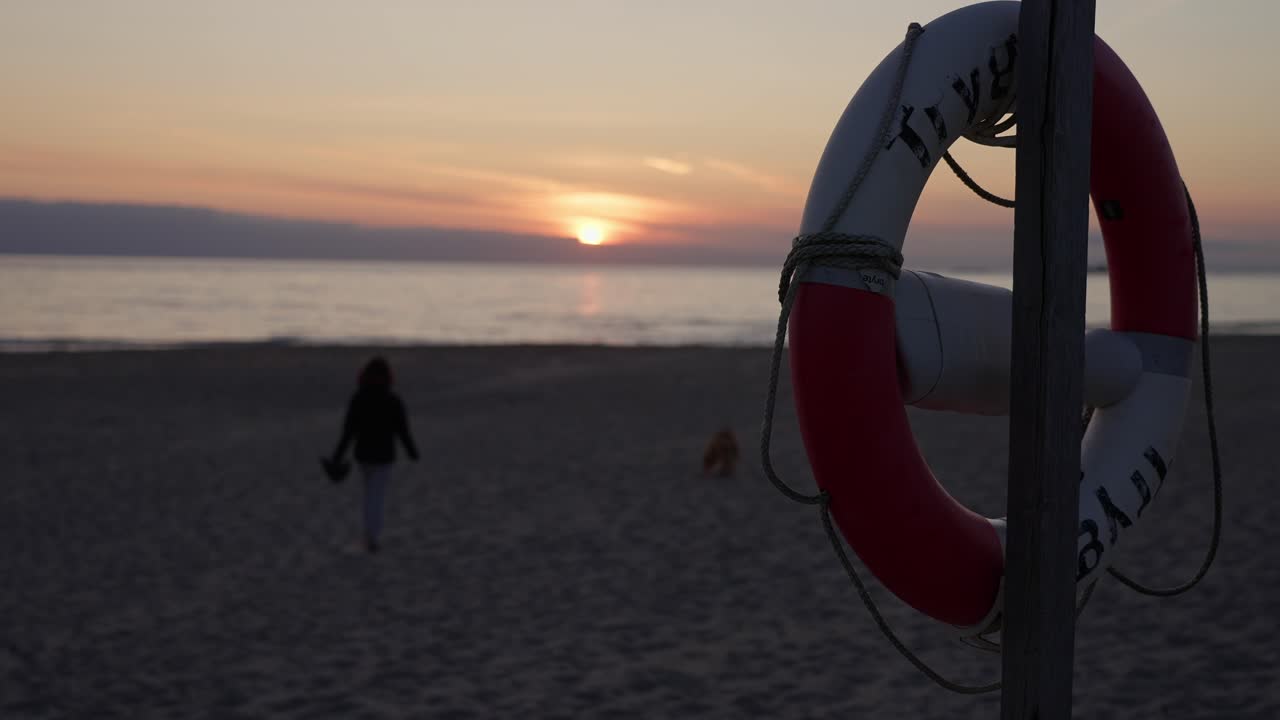 A woman walking along the beach with her dog at sunset, lifebuoy in foreground