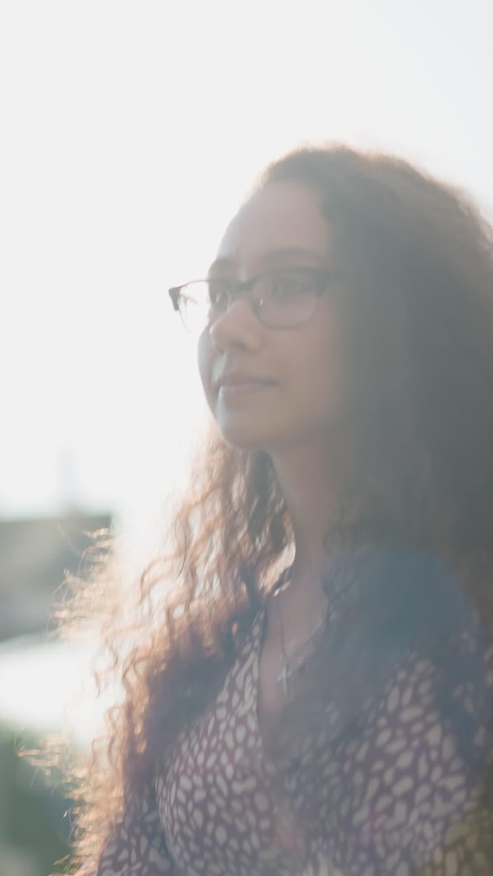 Young woman in glasses walks in busy city with a soft smile, glancing sideways as cars pass by, against background of urban buildings and soft sunlight