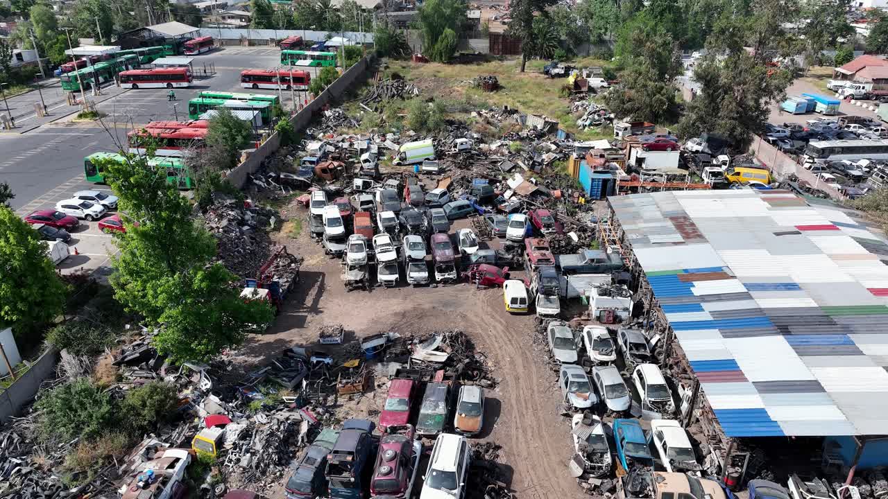 Forward drone aerial of junkyard with organized rows of scrapped cars, buses, warehouse buildings, and auto salvage depot overview