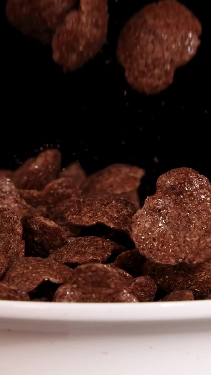 Fiocchi di cereali al cioccolato cadono a cascata in una ciotola di ceramica bianca su uno sfondo scuro da studio.