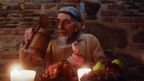 mature feudal lord has meal in medieval castle. elderly man guest opens wooden pint and drinks ale at dining table in king castle. dark ages period feast