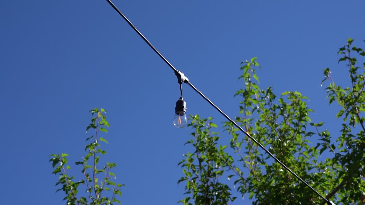Dim inline lightbulb lit on diagonal wire on blue sky summer day