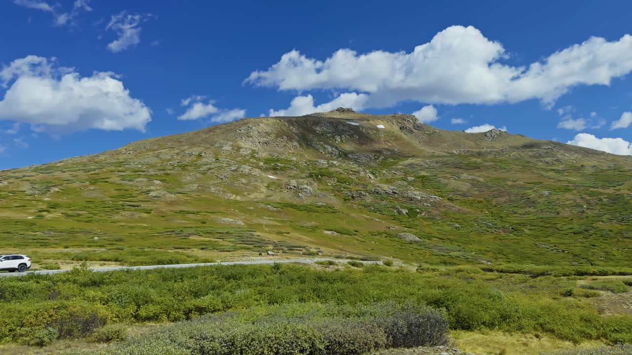 Sunny day at Independence Pass with rolling green terrain and scattered fluffy white clouds as SUV drives on dirt road