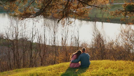 couple in love resting among autumn nature. young man and woman sitting on the hill and admiring the river at sunset.