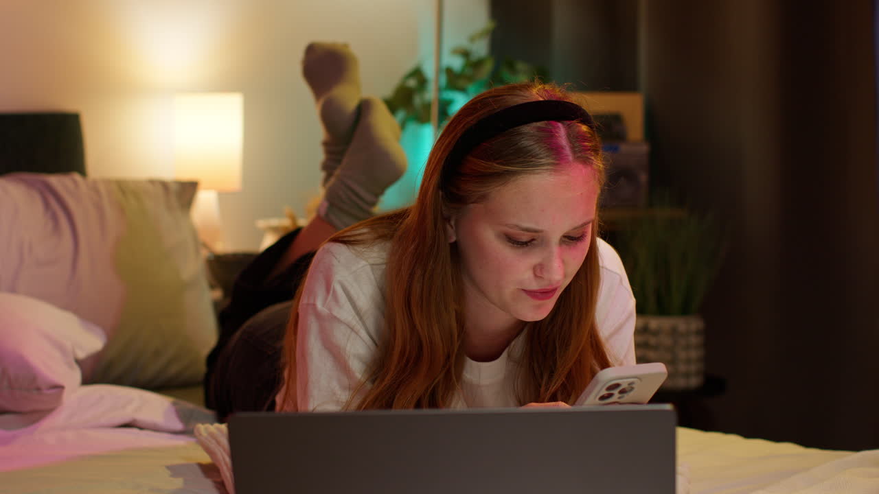 una mujer joven relajándose en la cama con una computadora portátil y un teléfono