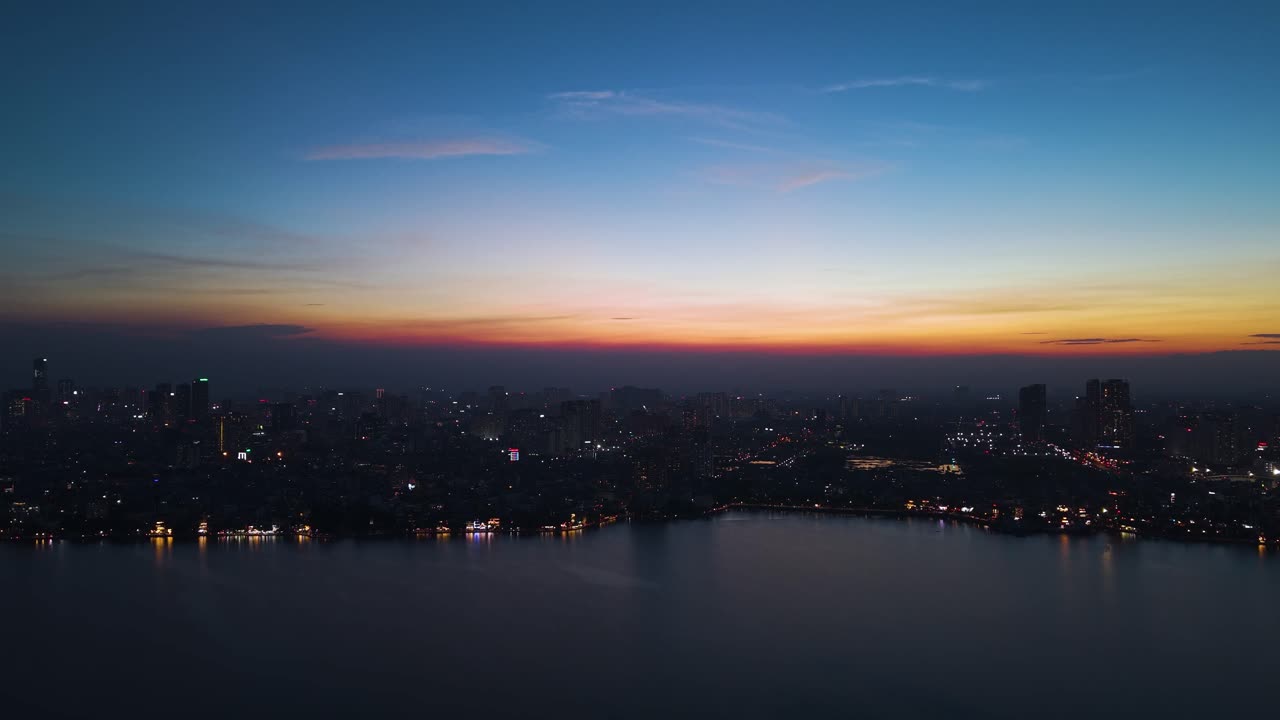 Sunset drone shot of Hanoi’s Tay Ho West Lake. Golden lights shimmer along the water’s edge, blending with the first hues of sunrise across the vibrant urban skyline