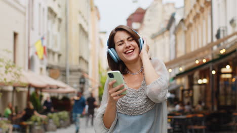 happy young woman in wireless headphones choosing, listening music dancing outdoors city street