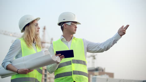 two colleagues of engineers a man and a woman discussing a drawing and a tablet computer on the background of buildings under construction and cranes a woman talking on the phone with the boss