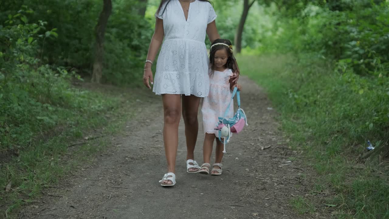 Una donna incinta e la sua giovane figlia, entrambe vestite di bianco, camminano mano nella mano attraverso un parco erboso. La scena cattura un momento sereno di legame familiare nella natura, circondate da alberi