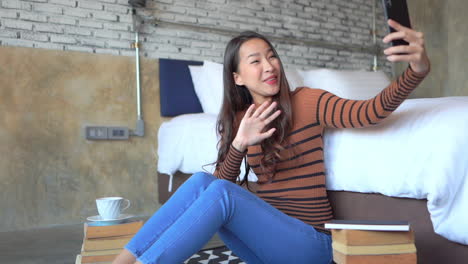 Young-female-Asian-student-with-long-dark-hair-sitting-on-floor-surrounded-by-book-waves-at-friend-or-family-on-video-call-using-smartphone