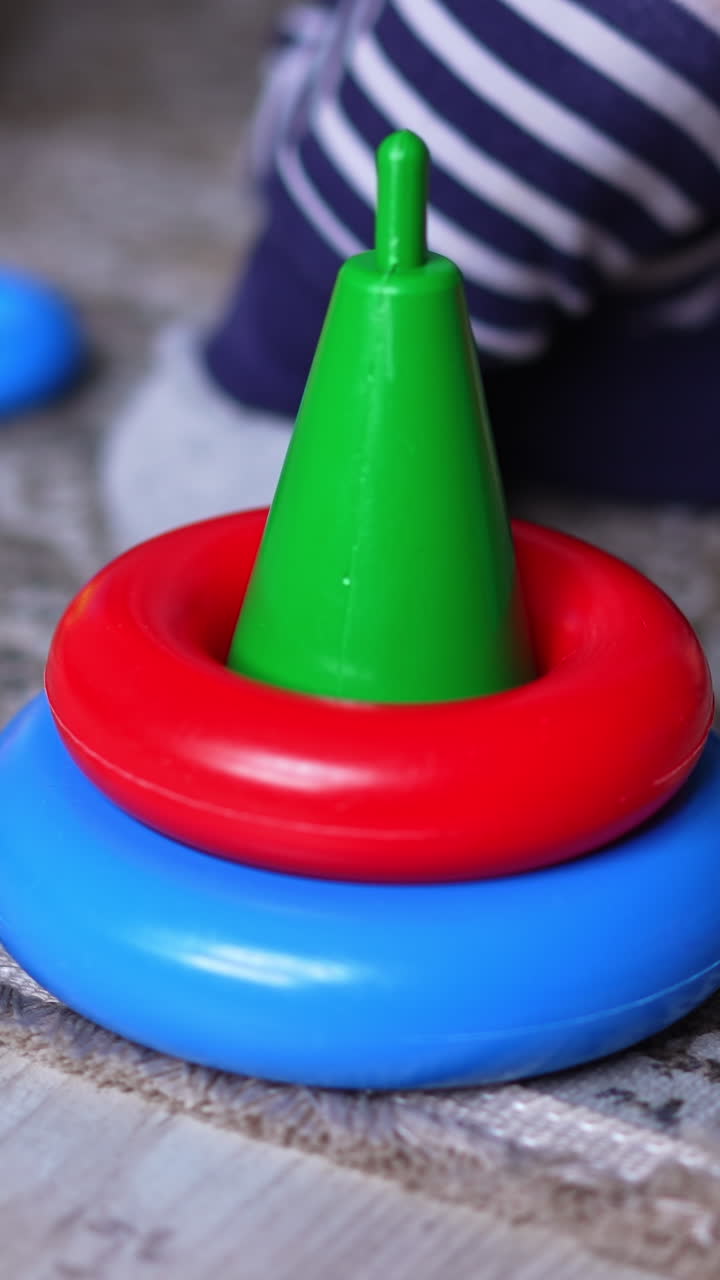 Little bright toy pyramid without upper parts. Close up. Unrecognized baby sitting on the floor next to a toy takes a red ring away. Blurred backdrop. Vertical video