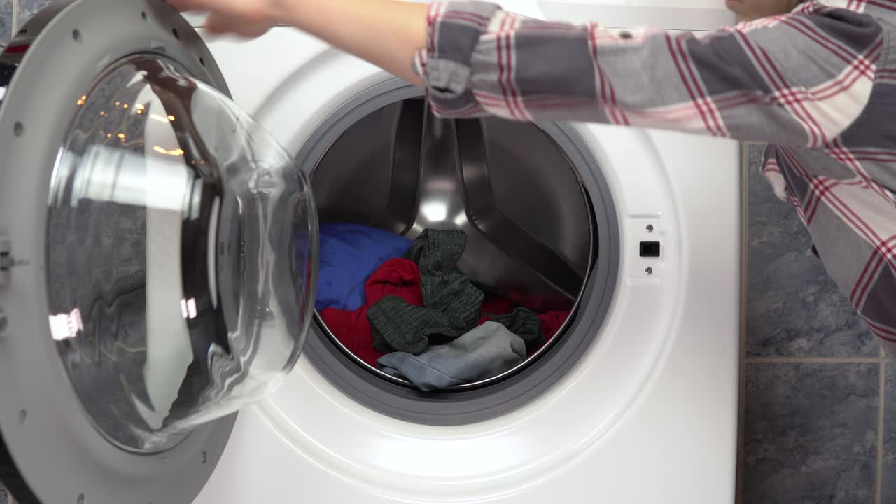 한 젊은 여성이 세탁기에서 세탁기를 열고 옷을 던지고 닫습니다.