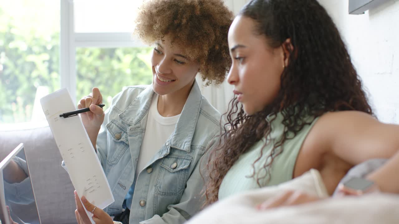 African American women left woman pointing with pen at paper, guiding partner reading on sofa