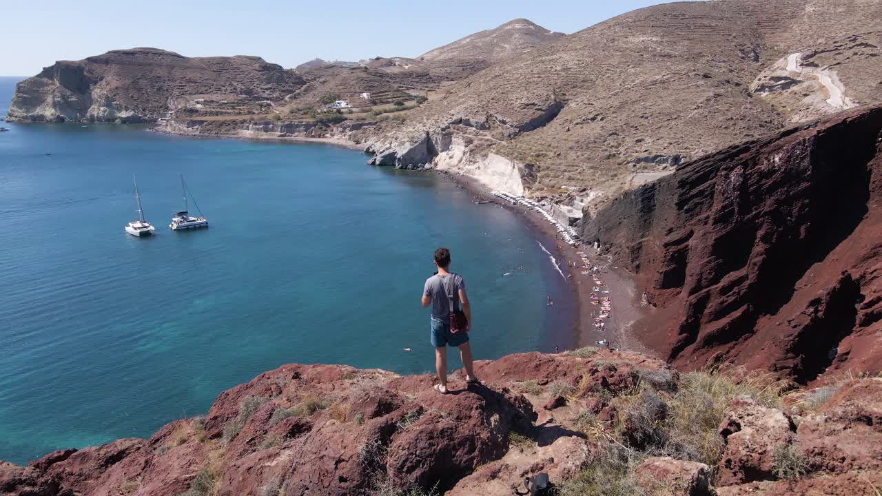 toma aérea sobre un hombre parado en el borde del acantilado sobre la playa roja en santorini, grecia