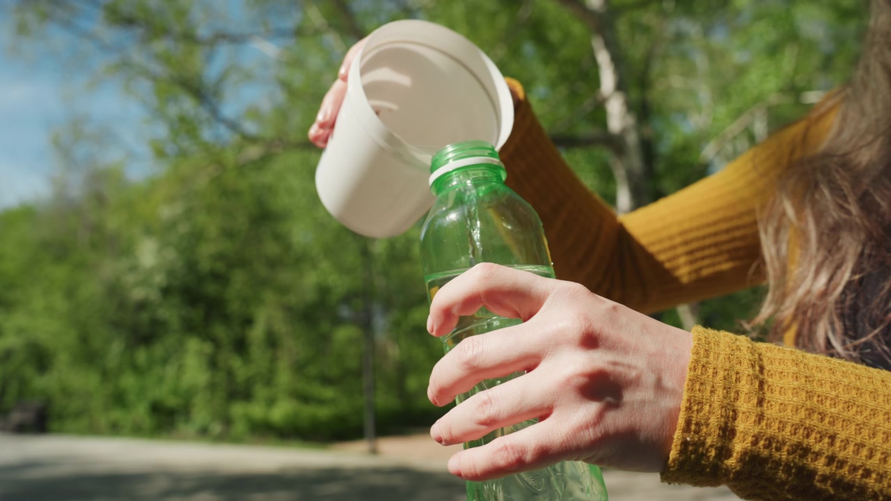 Una donna in giallo versa dell'acqua in una bottiglia, con una tavolozza di colori aperta e pennelli su una panchina del parco. La scena è ambientata all'aperto, con alberi verdi e luce solare che filtra.