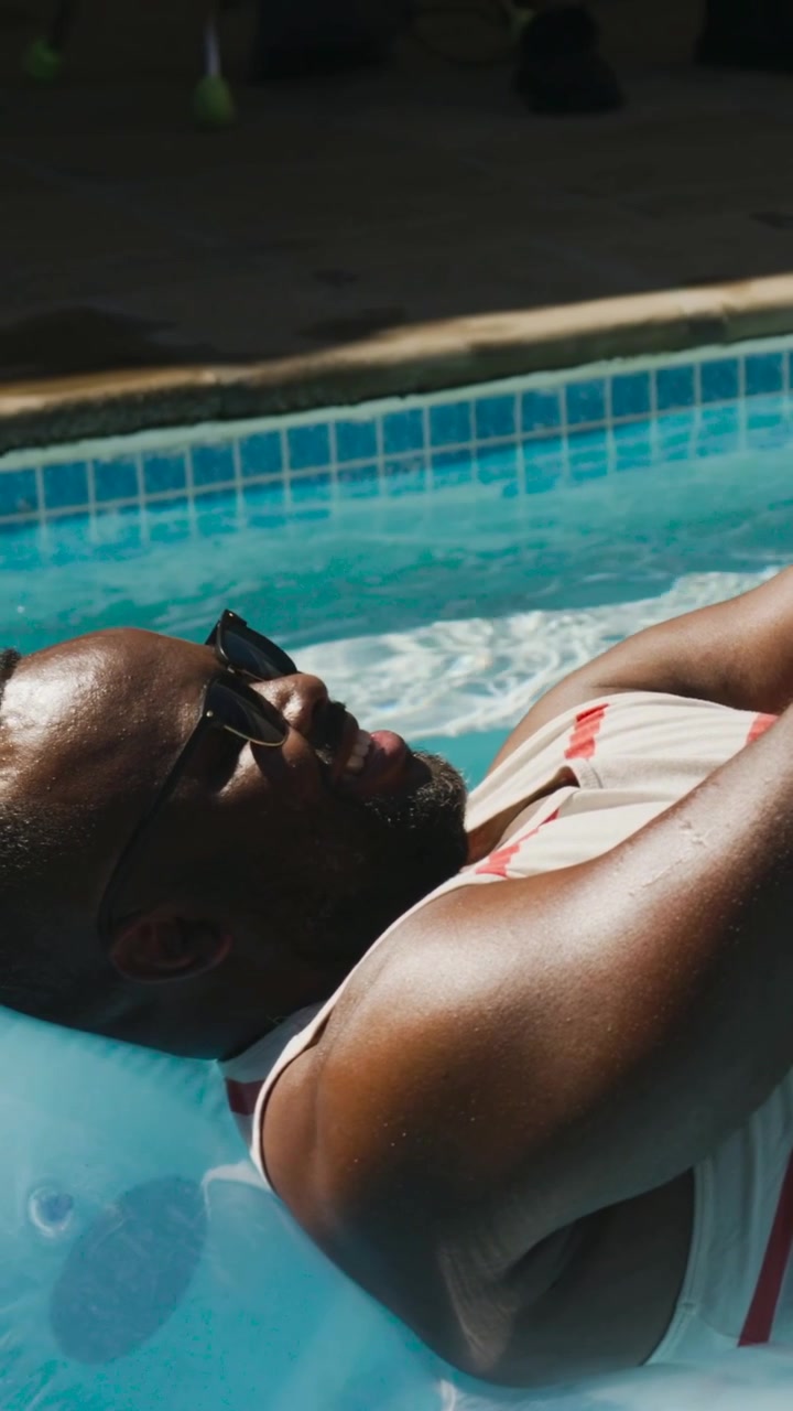 Vertical video: Drifting man shifting in pool after waves nudging float, in striped tank and shades