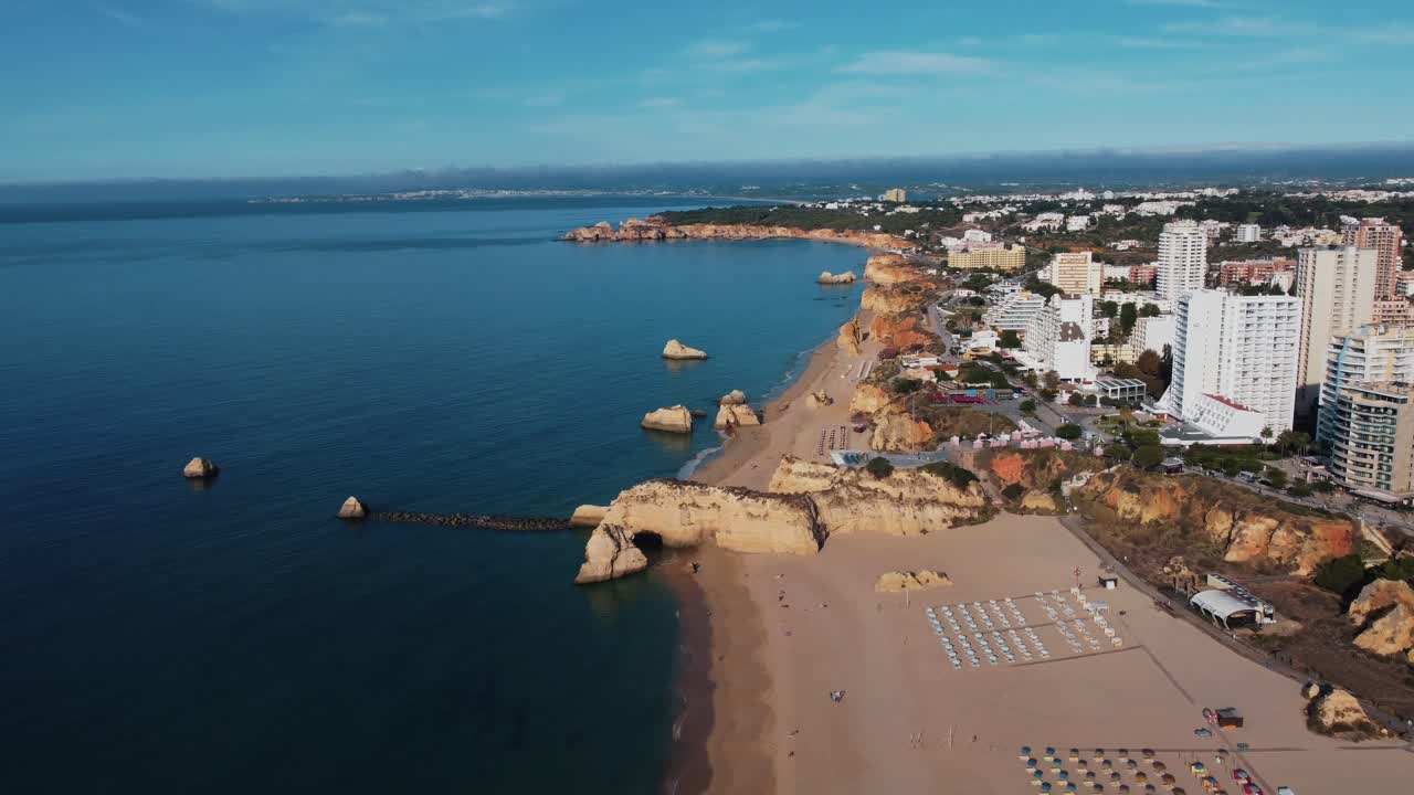 Drone view of Praia da Rocha with sandy beach, ocean waves, rock formations, and high-rise buildings along coastline under blue sky in Faro, Portugal showing mix of nature and urban scenery
