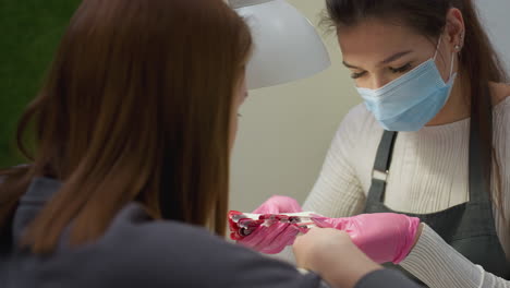nail technician wearing mask and pink gloves presents nail polish color samples to customer for selection during consultation at manicure station under lamp in modern beauty salon