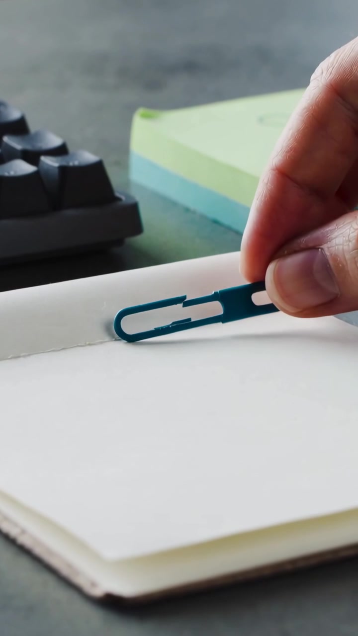 Video verticale: Mano che entra, allinea e posiziona una clip color verde acqua su un quaderno aperto sulla scrivania per segnare una pagina