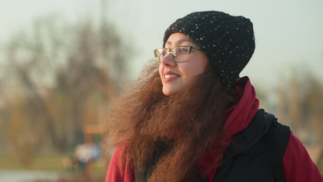 Young woman with glasses in park during golden hour, student pauses on a stroll, adjusts beanie and breathes cold air while smiling and gazing upward cozy red jacket and scarf, curly hair catching