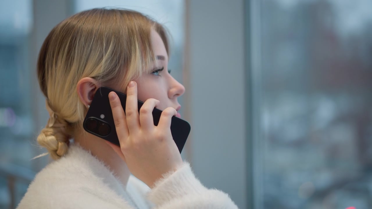 Waist-up profile of young woman holding smartphone to ear, speaking while lifting other hand to adjust hair beside bright window, with soft daylight, calm mood, and natural indoor setting