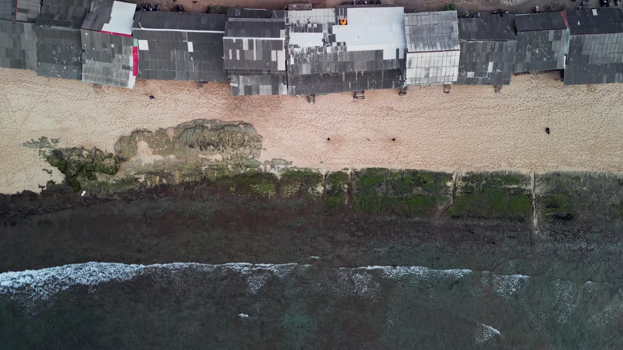 Overhead drone footage of Indonesian tropical coastline with white sand beachfront, low tide reef shelves, surf huts on the shore and steady ocean swell moving along the shallow reef edge