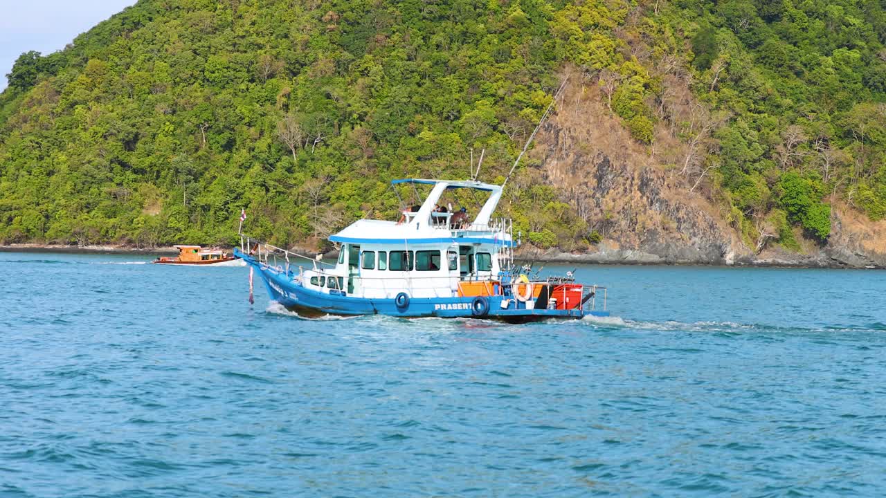 A fishing boat navigates calm waters near a lush, green island in Phuket, Thailand. Bright daylight enhances the serene ocean scene