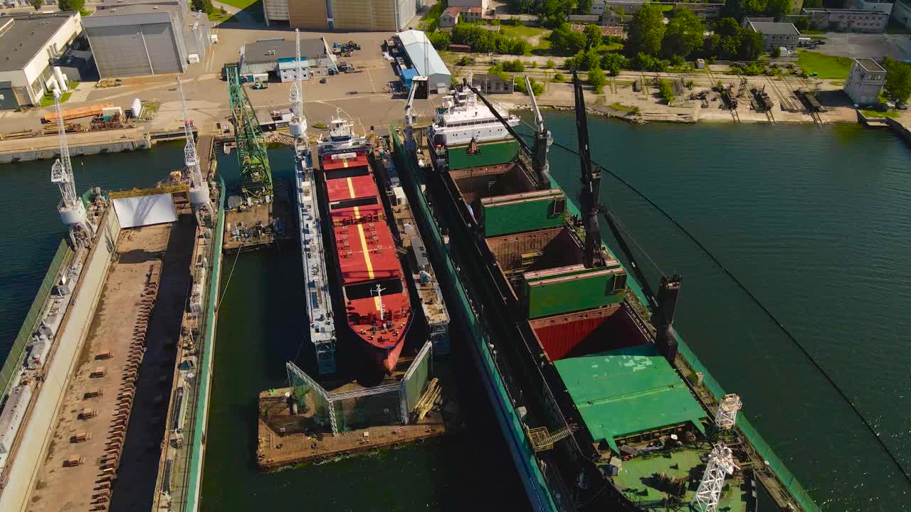 Aerial drone video flying over a large metal ship that is under construction at a harbor port during sunny summer day. The ship is raised above sea level and has large metal cranes around it working