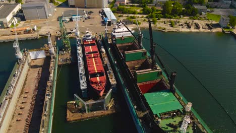 Aerial drone video flying over a large metal ship that is under construction at a harbor port during sunny summer day. The ship is raised above sea level and has large metal cranes around it working