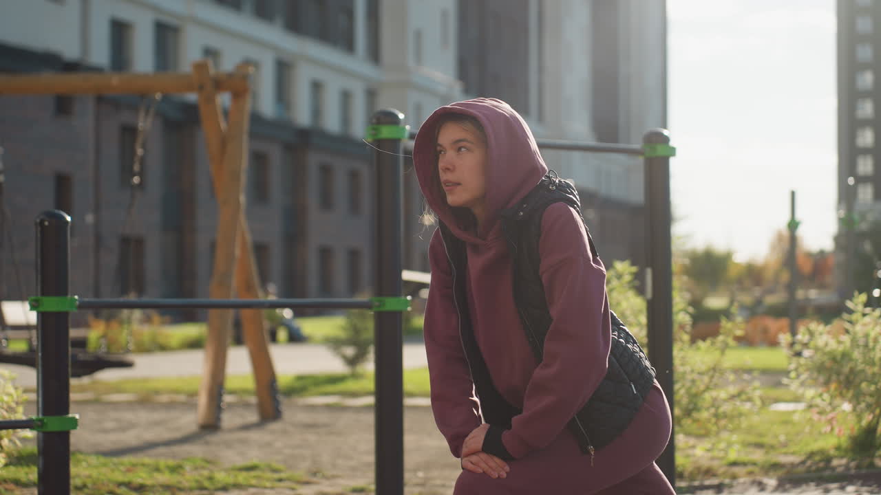 Energetic athlete in hoodie and quilted vest lunges forward under sunset glare in urban park with fitness bars, tall buildings backdrop and bystander, warming up muscles for dynamic training