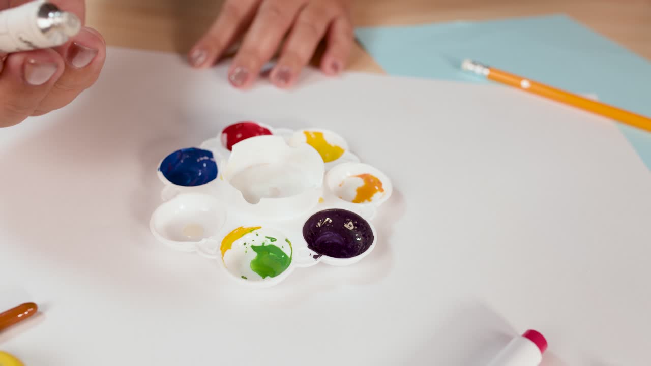 Child’s hand squeezes white paint into palette on desk, bright lighting, close-up, creative workspace
